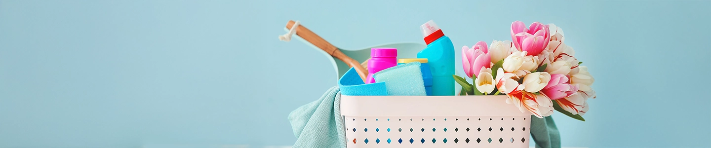 Ein weißer Korb mit Putzutensilien und rosa Tulpen vor hellblauem Hintergrund.