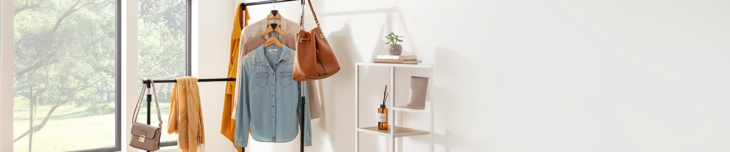 Kleiderständer mit drei verschiedenen Hemden, einem Schal, einem orangefarbenen Mantel und Handtaschen in der Nähe eines Fensters. Regale mit Dekoartikeln und einer Pflanze auf der rechten Seite.