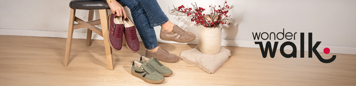 Eine Person sitzt auf einem Hocker und hält burgunderrote Turnschuhe in der Hand; auf dem Boden liegen beige und grüne Turnschuhe neben einer Vase mit roten Stielen.