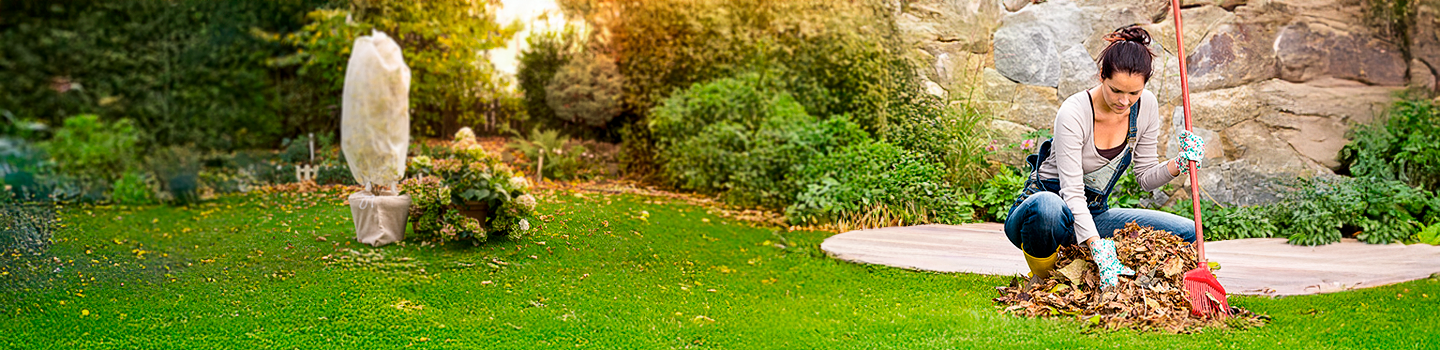 Eine Frau harkt Blätter in einem Garten, sie hockt auf einem Rasen mit einer Steinskulptur und üppigem Grün im Hintergrund.