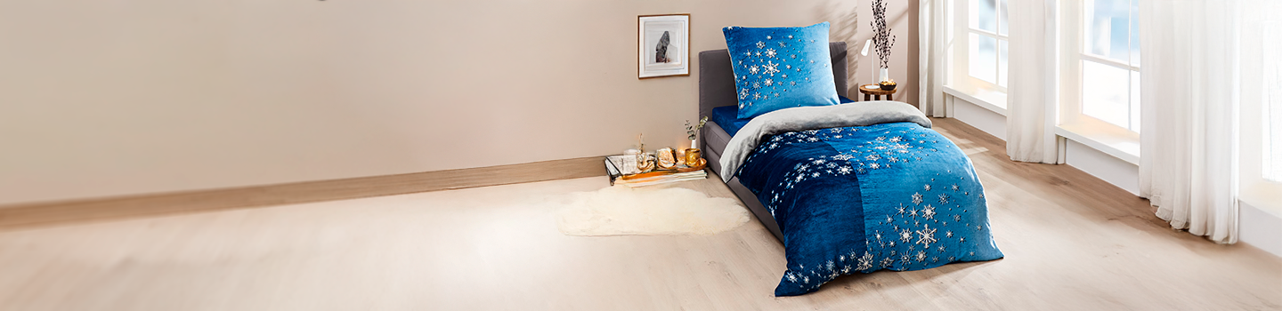 Gemütliches Schlafzimmer mit einem Bett mit blauer Bettwäsche mit Schneeflockenmuster, weichen Kissen und hellem Tageslicht durch große Fenster.