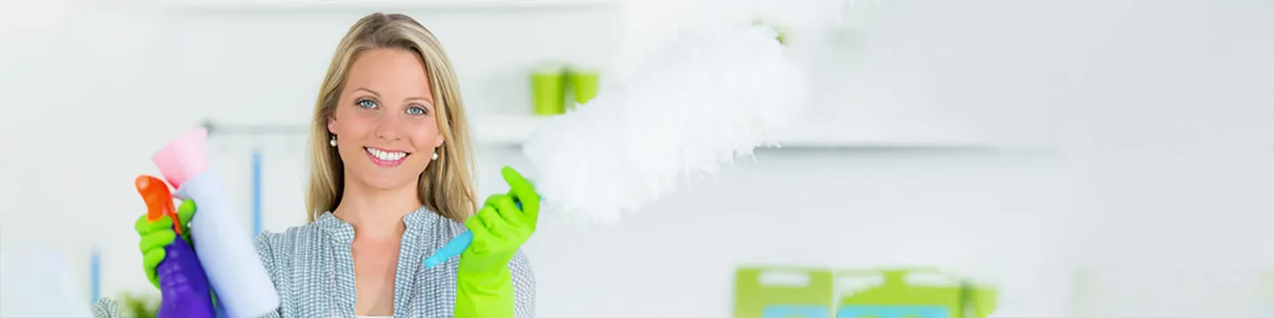 Eine lächelnde Frau hält Reinigungsmittel in einem hellen Raum; sie trägt grüne Handschuhe und ein graues Oberteil.