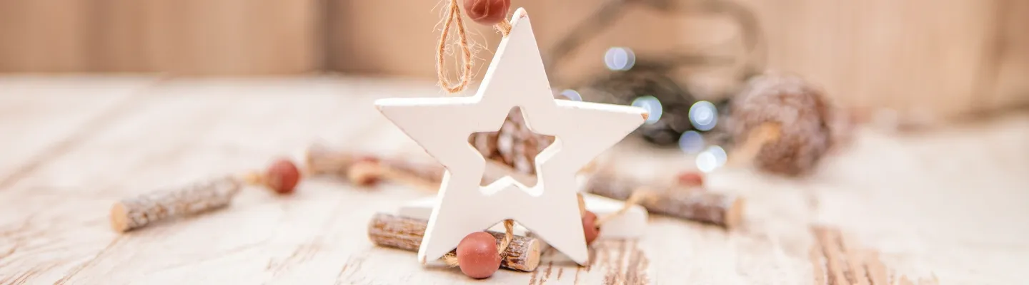 Ein weißes, sternförmiges Ornament mit einem ausgeschnittenen Stern hängt auf einer Holzoberfläche, umgeben von kleinen Holzscheiten und roten Beeren.