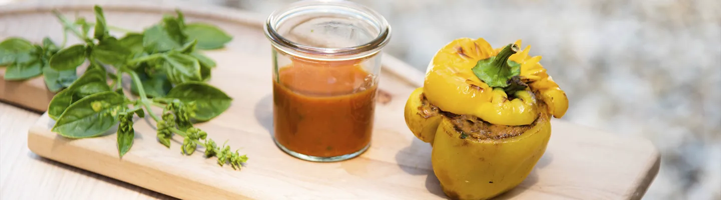 Gefüllte gelbe Paprika auf einem Holzbrett, dazu ein Glas Tomatensoße und frische Basilikumblätter.