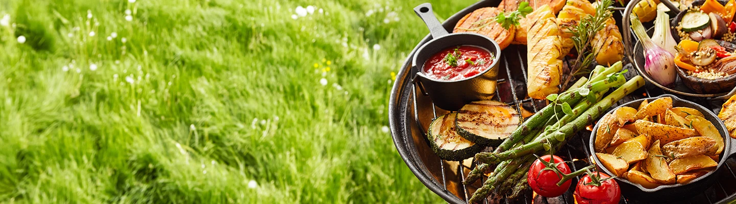 Ein Grill mit verschiedenen Gemüsesorten, Kartoffeln und Soße vor einer Kulisse aus saftig grünem Gras.