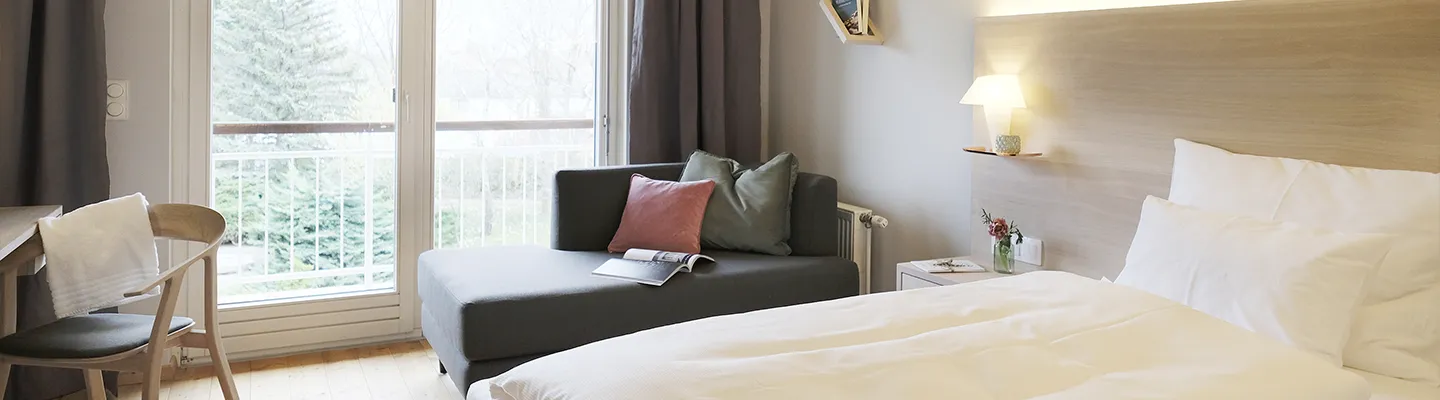 Helles, modernes Hotelzimmer mit einem gemütlichen Bett, einem grauen Sofa, einem kleinen Tisch und einem großen Fenster mit Blick ins Grüne. Sanfte Beleuchtung und minimalistische Einrichtung.