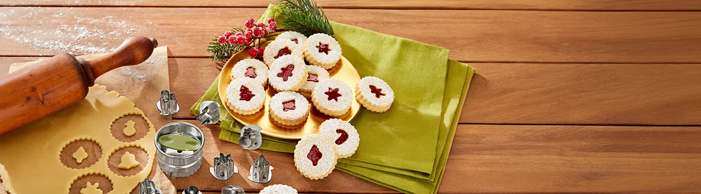 Ein Holztisch mit ausgestochenen Keksen und Teig, einem Nudelholz, Ausstechformen sowie einem gelben Teller mit Keksen mit Marmeladenfüllung auf einer grünen Serviette.