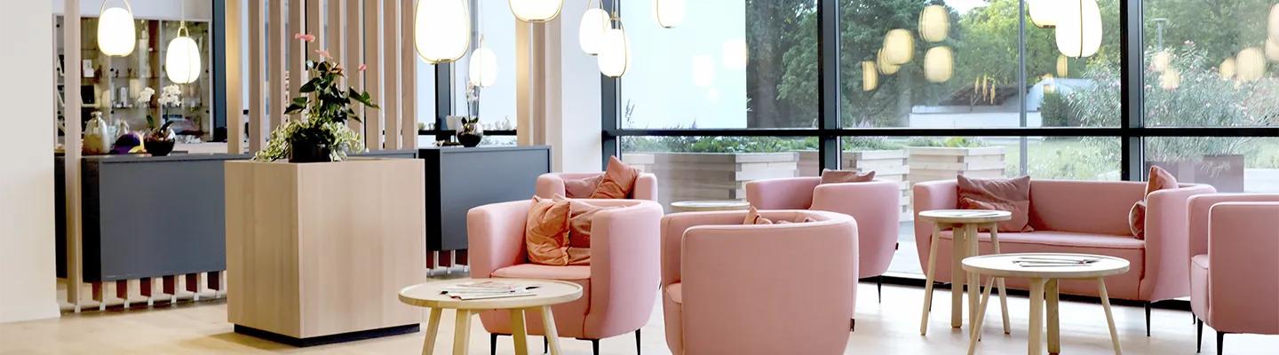 Moderne Lounge mit rosa Stühlen, runden Tischen und Pendelleuchten. Große Fenster bieten einen Blick auf den Garten und schaffen eine helle und einladende Atmosphäre.