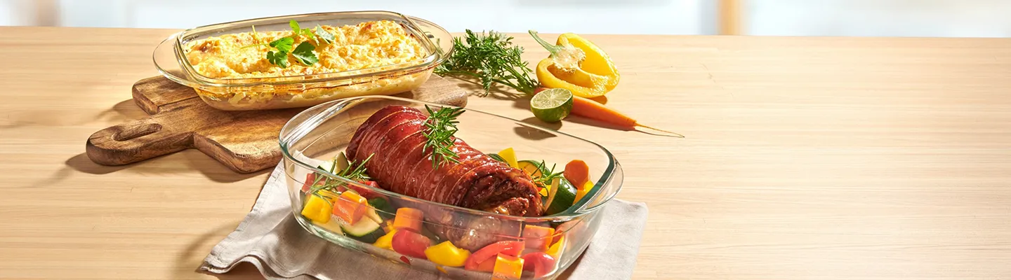 Roastbeef und Gemüse in einer Glasform neben einem überbackenen Auflauf, garniert mit Kräutern, auf einem Holztisch mit in Streifen geschnittener Paprika und Limette.