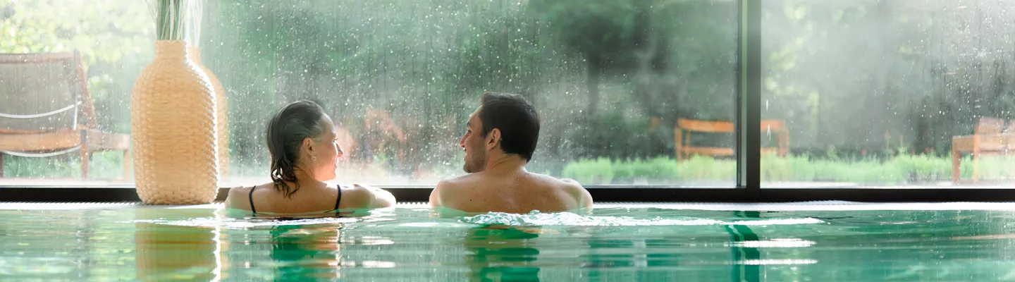 Ein Paar entspannt sich in einem Innenpool, schaut sich dabei an, während durch ein großes Fenster ein verschwommener Blick auf die grüne Landschaft draußen zu sehen ist.
