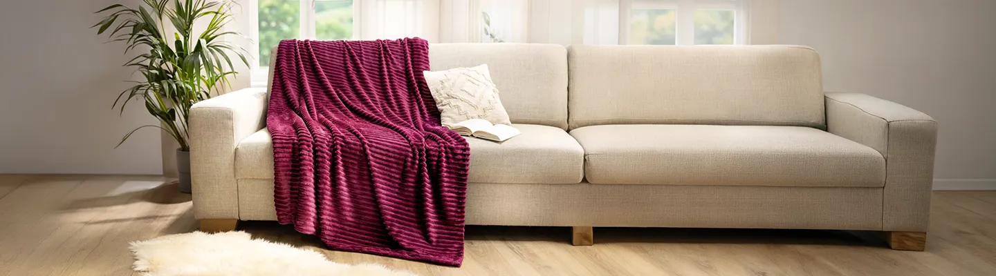 Ein beiges Sofa mit einer violetten Decke mit Strukturmuster und einem cremefarbenen Kissen, aufgestellt in einem hellen Raum mit Holzböden und einer Topfpflanze in der Nähe.