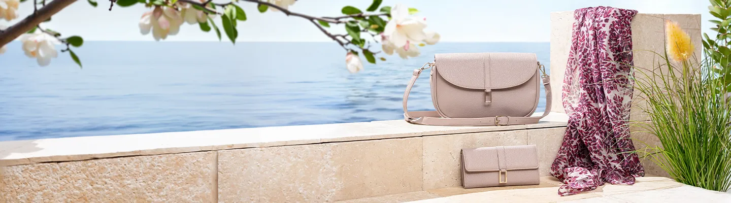 Eine beige Handtasche und eine Geldbörse sowie ein geblümter Schal liegen auf einem Felsvorsprung am Meer, umgeben von Grün und blühenden Blumen.
