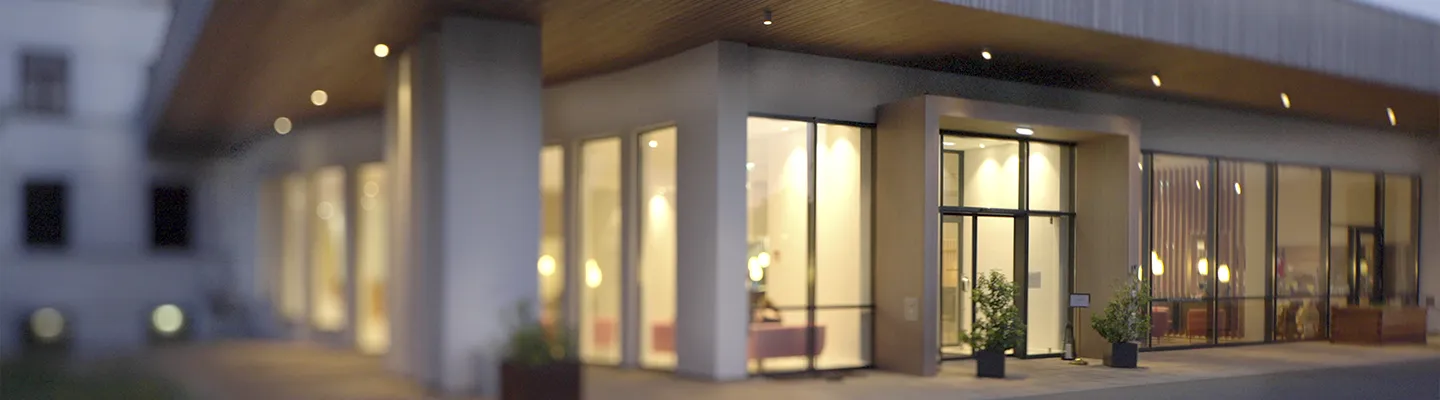 Moderner Gebäudeeingang mit großen Glasfenstern, warmer Innenbeleuchtung und Topfpflanzen im Außenbereich.