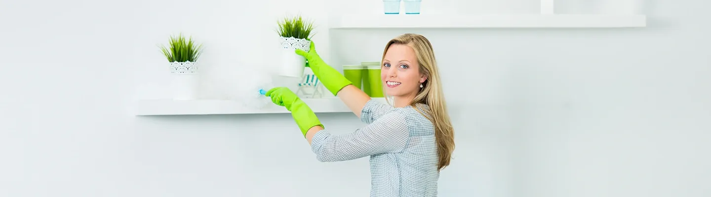 Eine Frau trägt grüne Handschuhe und staubt ein weißes Regal mit Topfpflanzen ab, während sie in die Kamera lächelt.