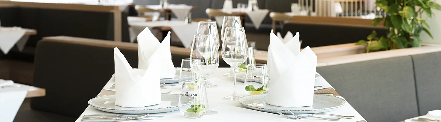 Elegantes Restaurant mit weißer Tischdecke, gefalteten weißen Servietten, Weingläsern und Besteck, im Hintergrund moderne Sitzgelegenheiten.