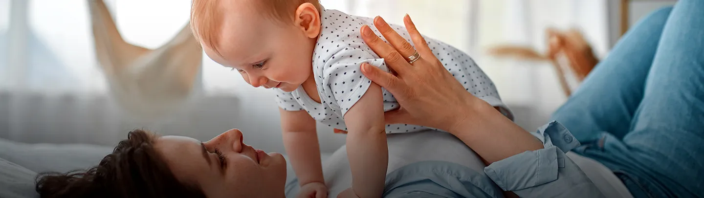 Eine Person liegt auf einem Bett und lächelt ein Baby in einem gepunkteten Outfit an, das spielerisch auf ihrer Brust in einem sonnenbeschienenen Raum liegt.