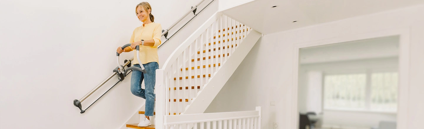 Eine Frau in einem gelben Pullover benutzt einen Treppenlift auf einer weißen Treppe in einer hellen, modernen Wohnung.