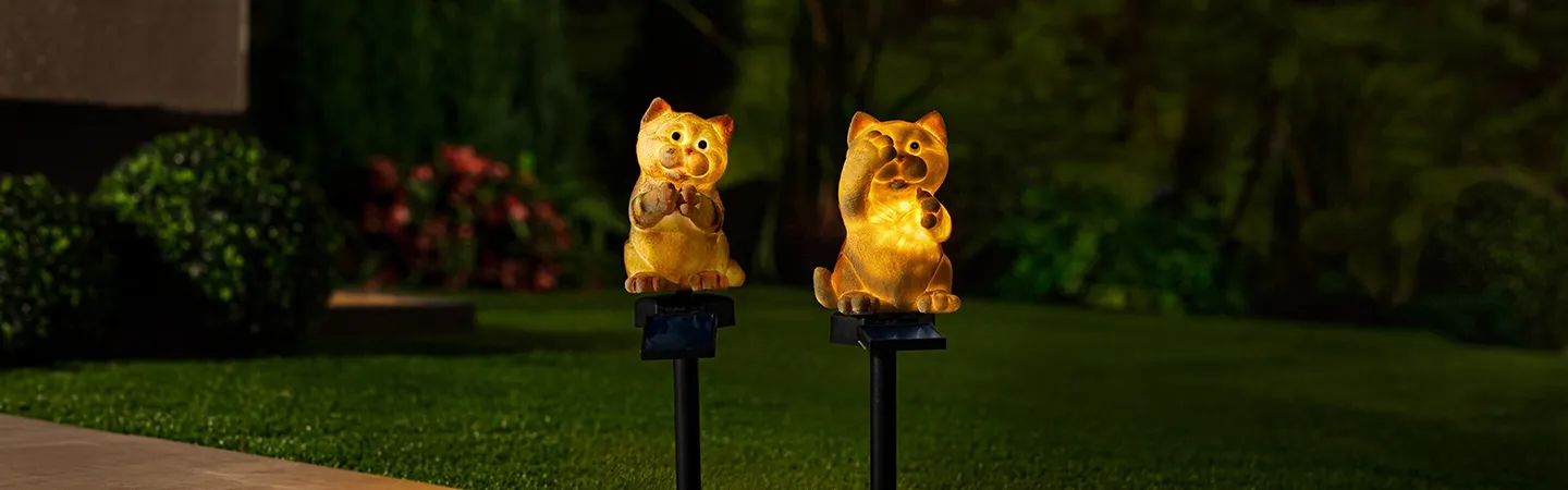 Zwei Solarleuchten in Katzenform auf Erdspießen beleuchten nachts einen Garten, umgeben von Pflanzen und einem dunklen Hintergrund.