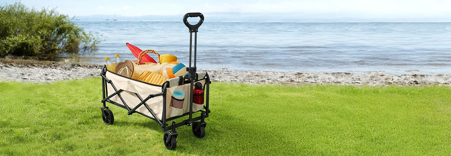 Ein Strandwagen, gefüllt mit buntem Strandzubehör wie einem Hut, einem Ball und Handtüchern, steht auf Gras nahe einer felsigen Küstenlinie mit ruhigem Wasser.