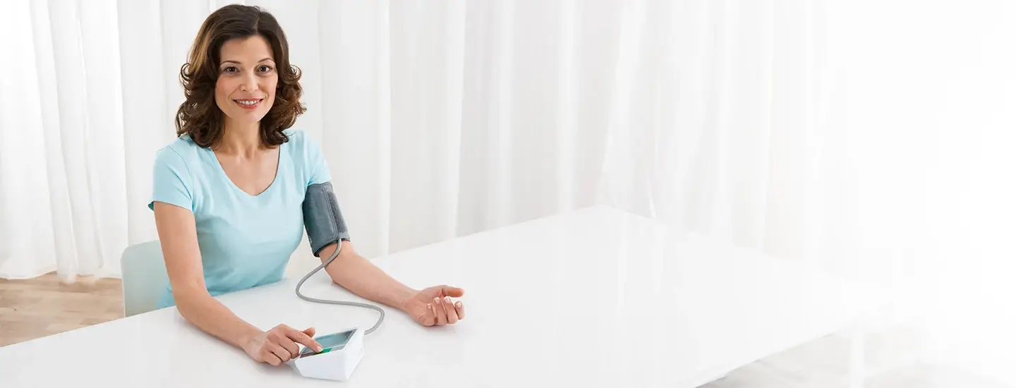 Frau, die ihren Blutdruck mit einem digitalen Messgerät überprüft, während sie an einem weißen Tisch in einem hellen Raum mit transparenten Vorhängen sitzt.