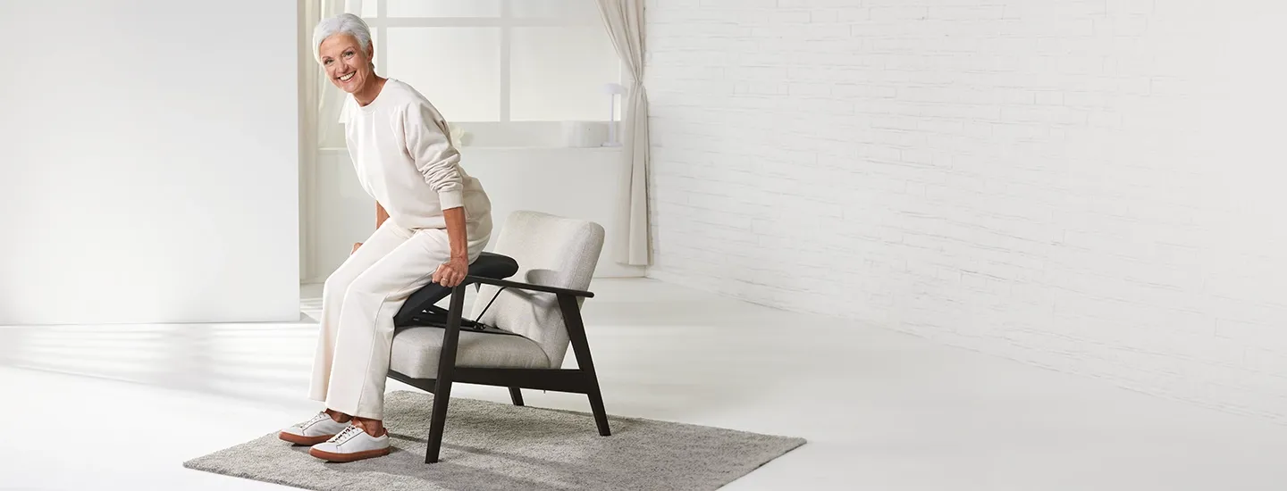 Eine lächelnde ältere Frau in weißer Kleidung steht in einem hellen, minimalistisch eingerichteten Raum von einem modernen Sessel auf.