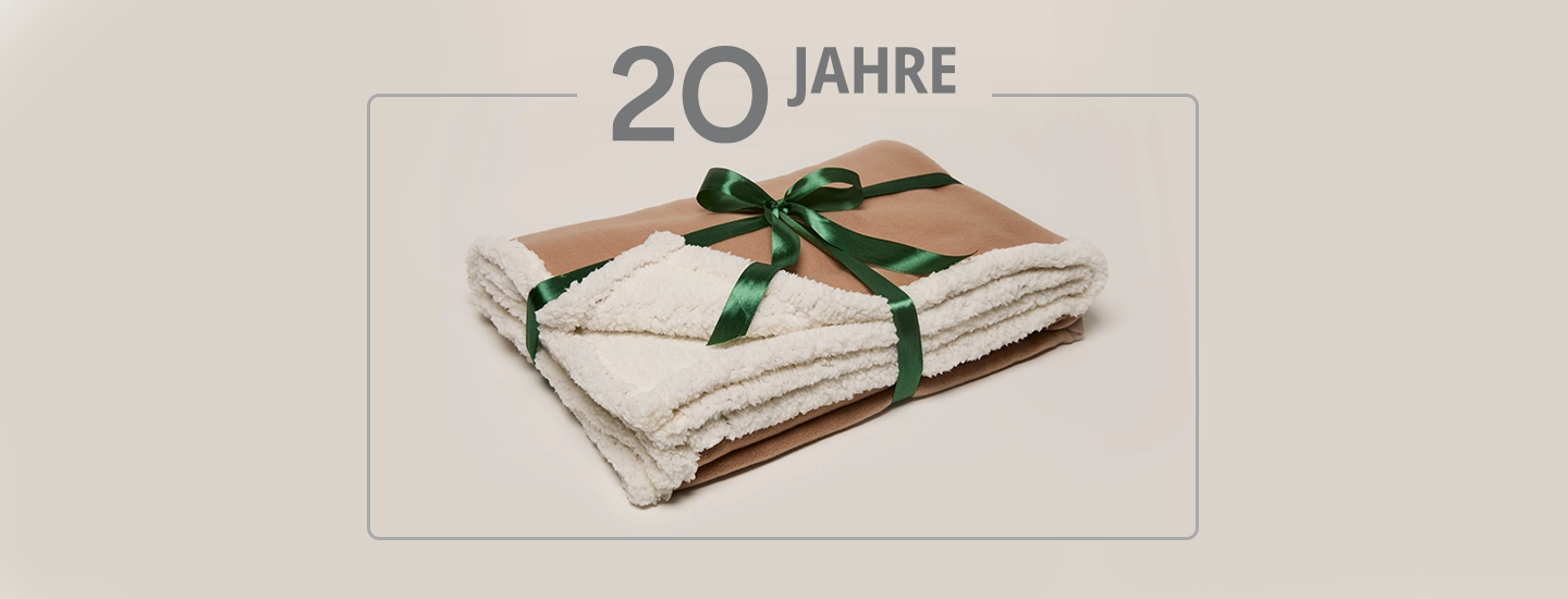 Eine gefaltete beige Decke mit weißem, flauschigem Futter, zusammengebunden mit einem grünen Band. Der Text darüber lautet „20 Jahre“.