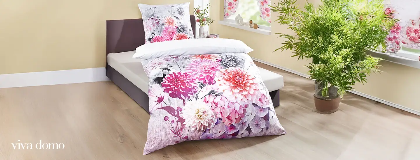 Bett mit Bettwäsche mit Blumenmuster in einem hellen Zimmer, mit großen Blumen in Rosa und Weiß. Helle Holzböden und eine Blattpflanze in der Nähe.