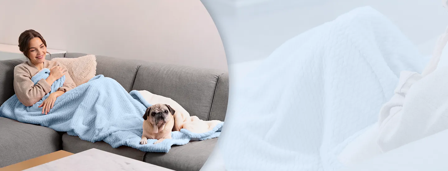 Eine Frau in einem beigen Pullover sitzt lächelnd auf einem grauen Sofa unter einer hellblauen Decke, neben ihr liegt ein Mops.