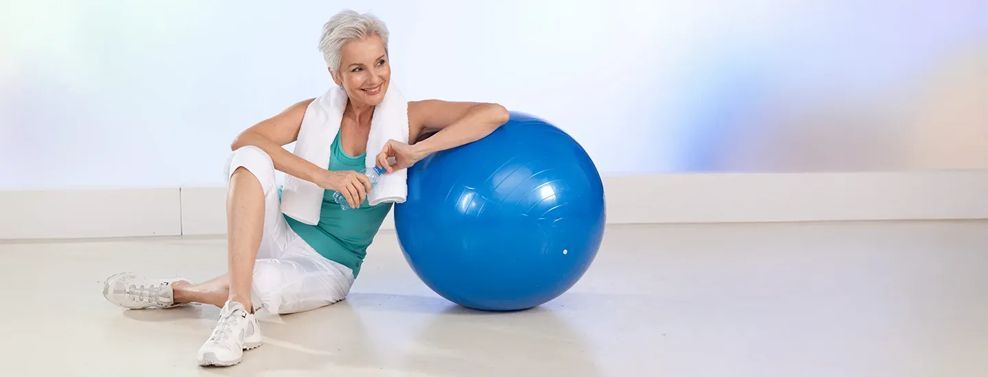 Eine lächelnde Frau mit kurzen grauen Haaren ruht auf einem blauen Gymnastikball, hält eine Wasserflasche in der Hand, trägt Sportkleidung und hat ein Handtuch über der Schulter.