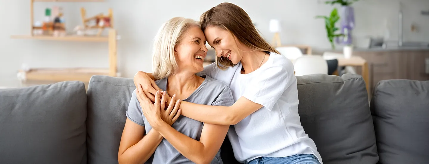 A smiling older woman and a younger woman embrace joyfully on a sofa in a bright living room.