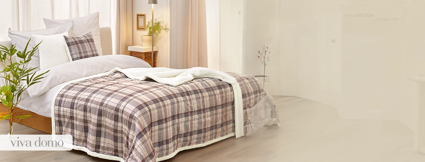 Gemütliches Schlafzimmer mit einer karierten Tagesdecke, weißen Kissen, einem Nachttisch mit Lampe und einer Pflanze. Sanftes Tageslicht erfüllt den Raum.