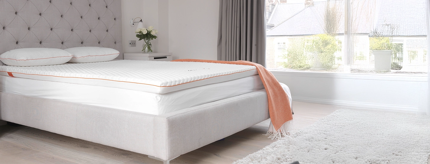 Ein helles Schlafzimmer mit einem ordentlich gemachten Bett, darauf ein weißer Matratzentopper, weiße Bettwäsche, eine orangefarbenen Tagesdecke, zwei Kissen. Daneben ein Beistelltisch mit Blumen.