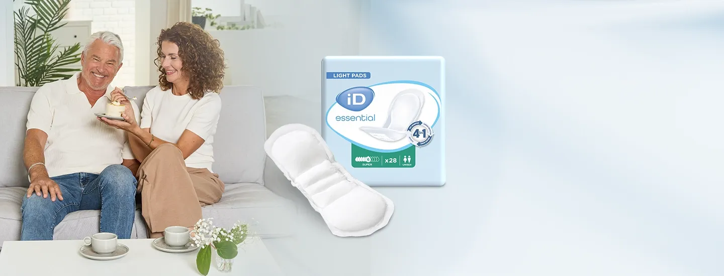 Ein älteres Ehepaar sitzt lächelnd mit einem Kuchen auf einem Sofa, daneben liegen ein Paket und einzelne „iD essential light pads“ vor einem hellblauen Hintergrund.