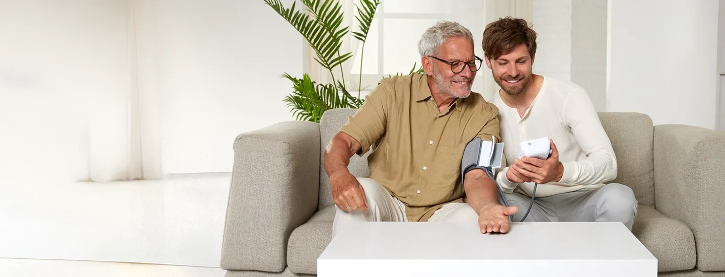 Blutdruckmessgerät mit Oberarmmanschette und digitaler Anzeige in Anwendung auf einer Person auf einem Sofa.