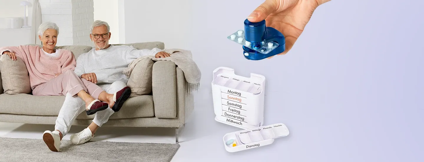 Blauer Tablettenteiler in einer Hand sowie weiße Wochendosierbox mit beschrifteten Tagesfächern; daneben sitzt ein älteres Paar auf einem hellen Sofa im Wohnbereich.