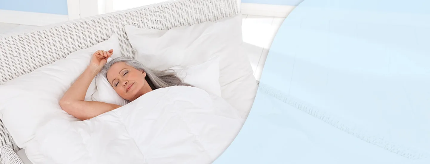 Eine Frau mit langen grauen Haaren schläft friedlich im Bett, zugedeckt mit einer weißen Decke, vor einem hellblauen und weißen Hintergrund.