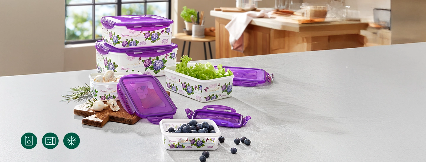 Verschiedene stapelbare, lila-weiße Vorratsbehälter mit Blumenmuster auf einer Küchenarbeitsplatte, gefüllt mit Beeren, Salat und Knoblauch.