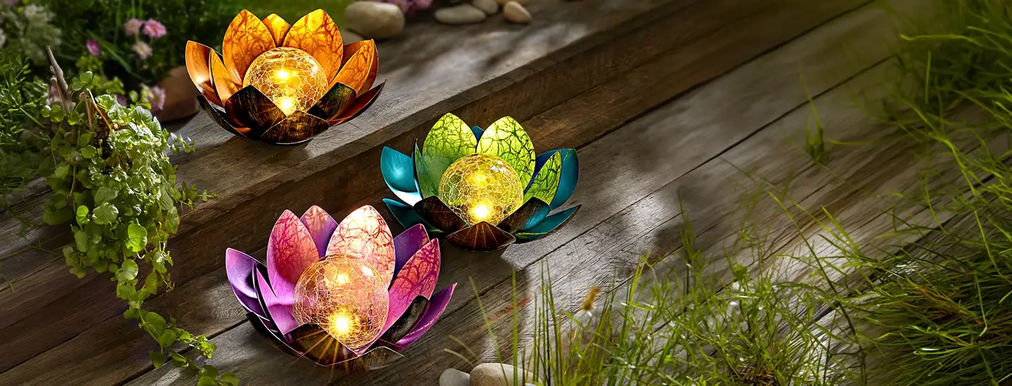 Drei farbenfrohe, lotusförmige Leuchten mit leuchtenden Glaskugeln, die auf Holzstufen in einer Gartenkulisse stehen, umgeben von Grünpflanzen und Felsen.