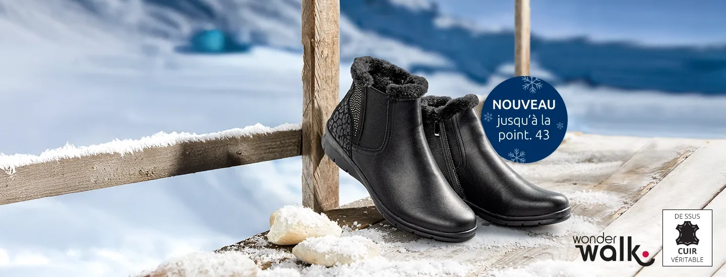 Schwarzes, knöchelhohes Paar Schuhe mit Reißverschluss, elastischem Einsatz und hellem Innenfutter auf einer schneebedeckten Holzfläche vor winterlicher Bergkulisse.