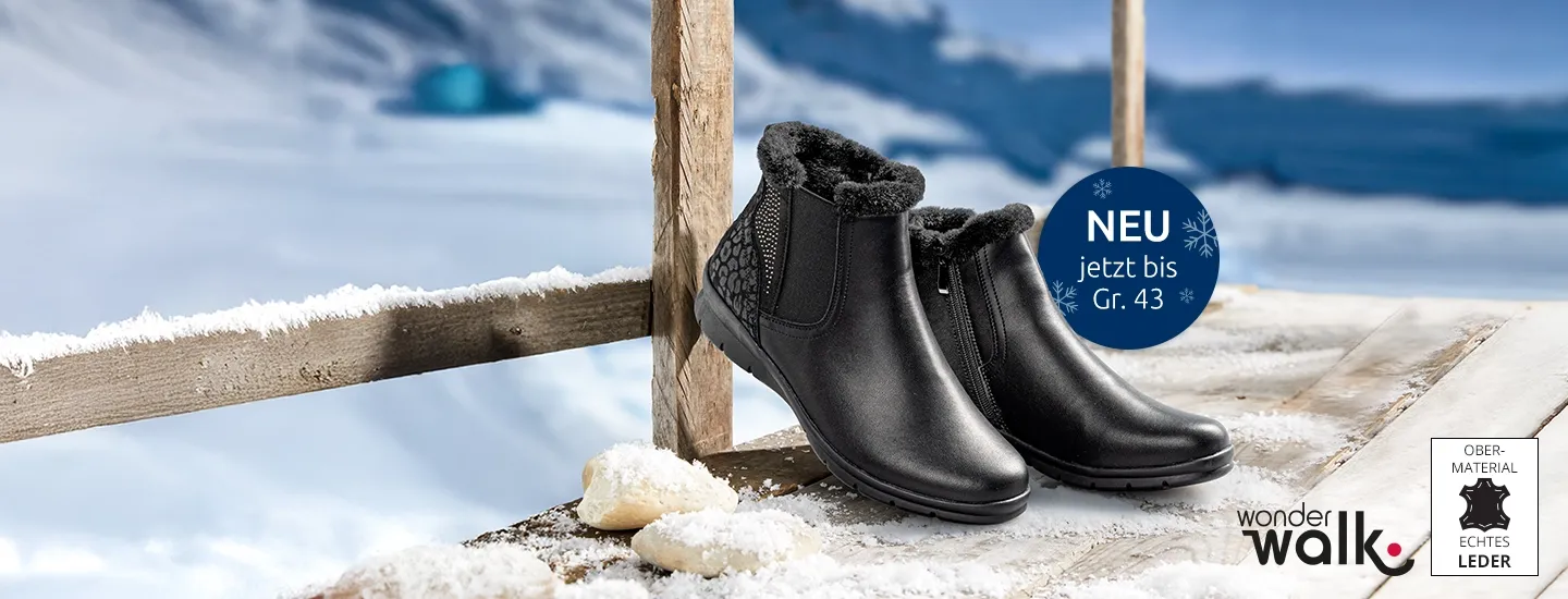 Schwarze Stiefeletten mit Fellfutter auf einer verschneiten Holzoberfläche vor der Kulisse einer winterlichen Schneelandschaft.