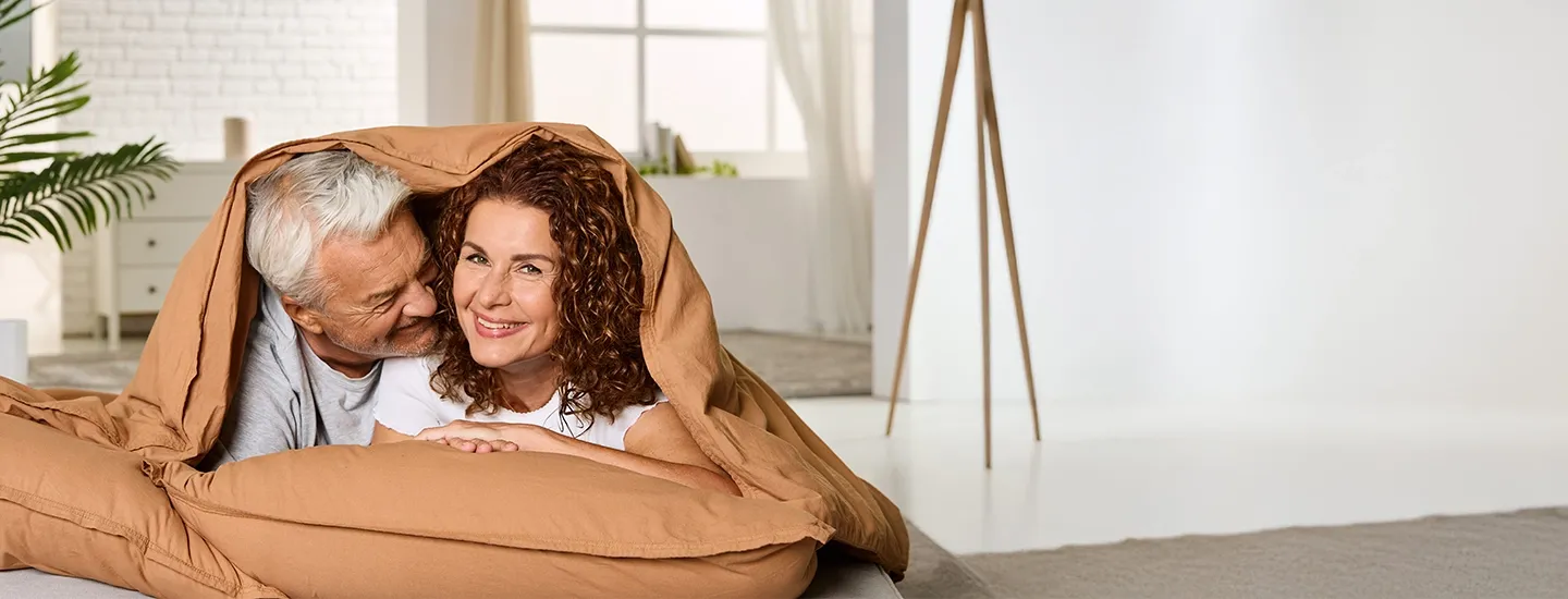 Ein älteres Paar, das lächelnd unter einer braunen Decke auf einem Bett in einem hellen, modernen Zimmer mit Pflanzen und einer Stehlampe kuschelt.