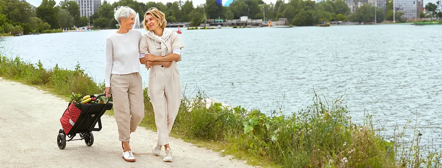 Zwei Frauen spazieren mit einem roten Einkaufswagen auf einem Weg am Seeufer entlang. Sie lächeln, sind leger gekleidet und von Grün und Wasser umgeben.