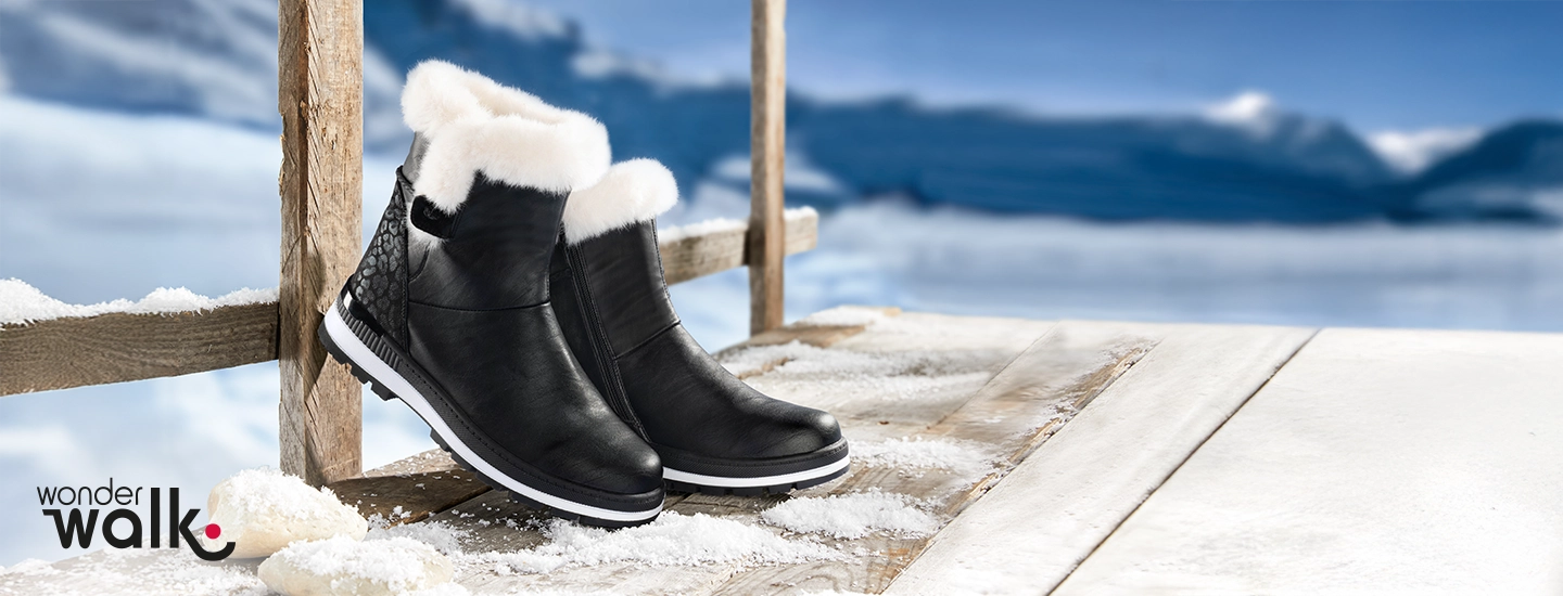 Schwarze Winterstiefel mit weißem Fellfutter stehen auf einem schneebedeckten Holzsteg, im Hintergrund eine verschneite Berglandschaft.