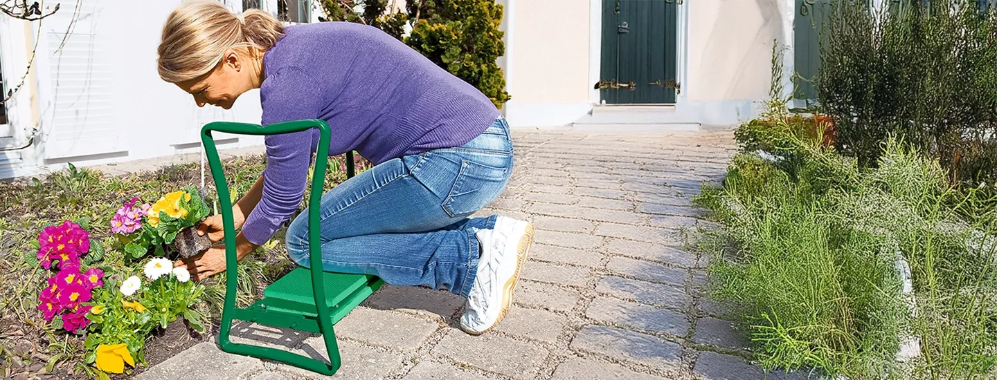 Eine Person in einem violetten Pullover kniet auf einer Gartenkniebank und kümmert sich um bunte Blumen entlang eines Steinwegs in einem sonnigen Garten.