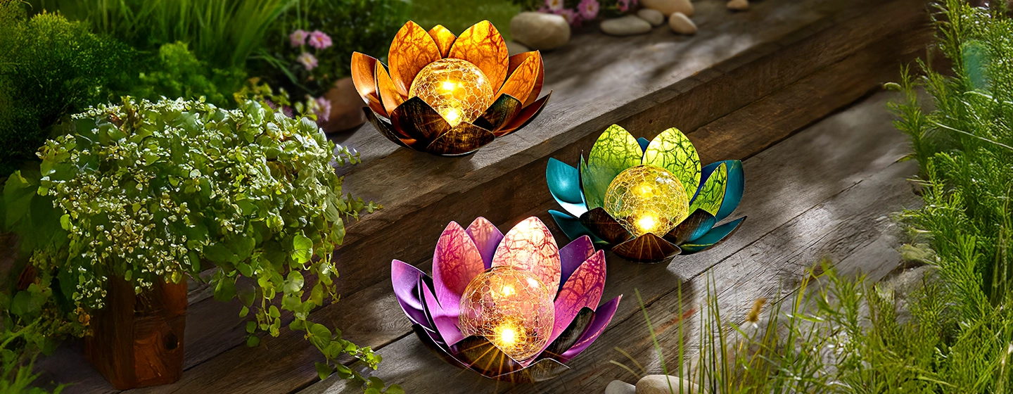 Drei farbenfrohe, lotusförmige Laternen, deren Mitte leuchtet, stehen auf einer Holzterrasse, umgeben von üppigem Grün und kleinen Blumen.