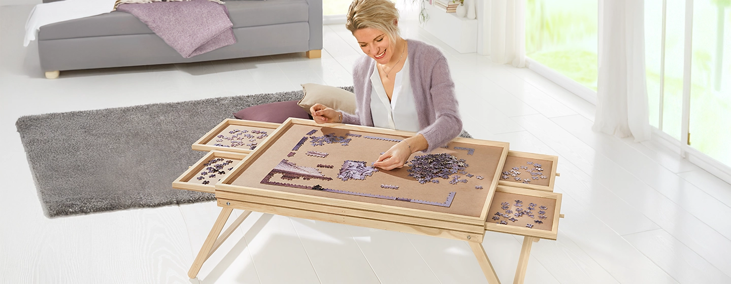 Frau, die auf einem Holztisch mit Schiebefächern in einem hellen Raum mit Sofa und großen Fenstern ein Puzzle zusammenbaut.