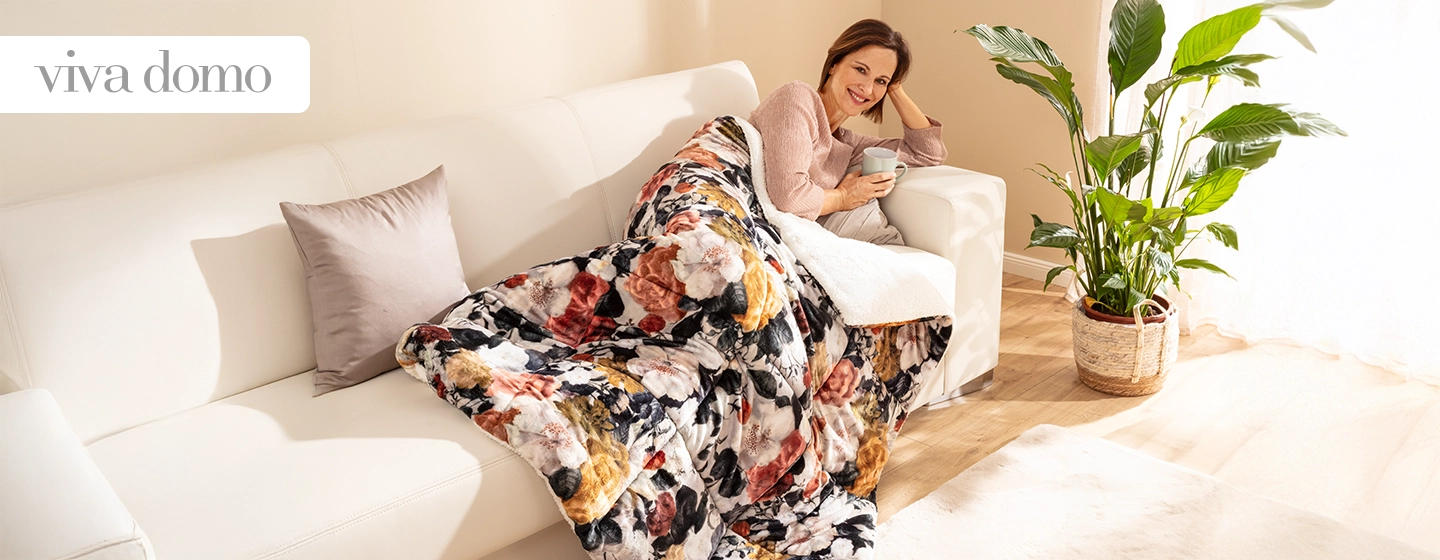 Frau, die sich auf einem weißen Sofa mit einer geblümten Decke entspannt und eine Tasse hält. In der Nähe steht eine große Topfpflanze, und Sonnenlicht erfüllt den Raum.