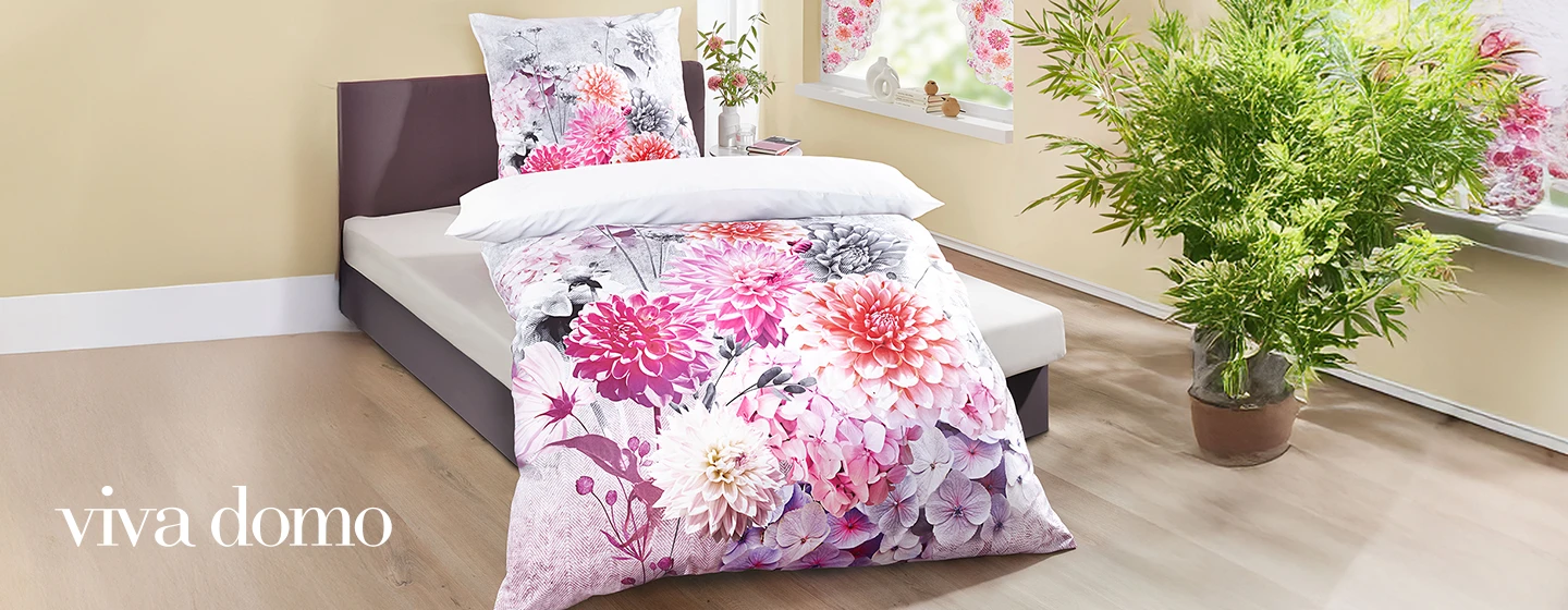 Ein Schlafzimmer mit einem Bett mit Bettwäsche und Kissen mit Blumenmuster, einer Topfpflanze und Fenstern mit Blumenvorhängen. Der Text lautet „viva domo”.