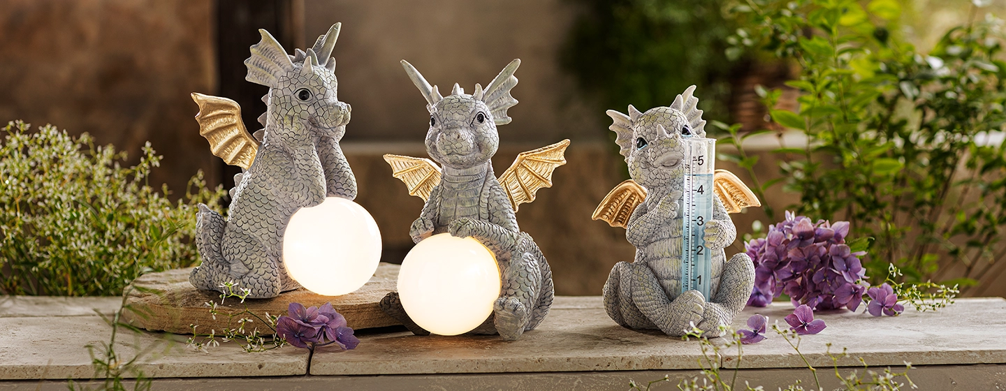 Drei dekorative Drachenstatuen auf einer Holzoberfläche, zwei davon halten Lichter und eine ein Thermometer, umgeben von Pflanzen und Blumen.