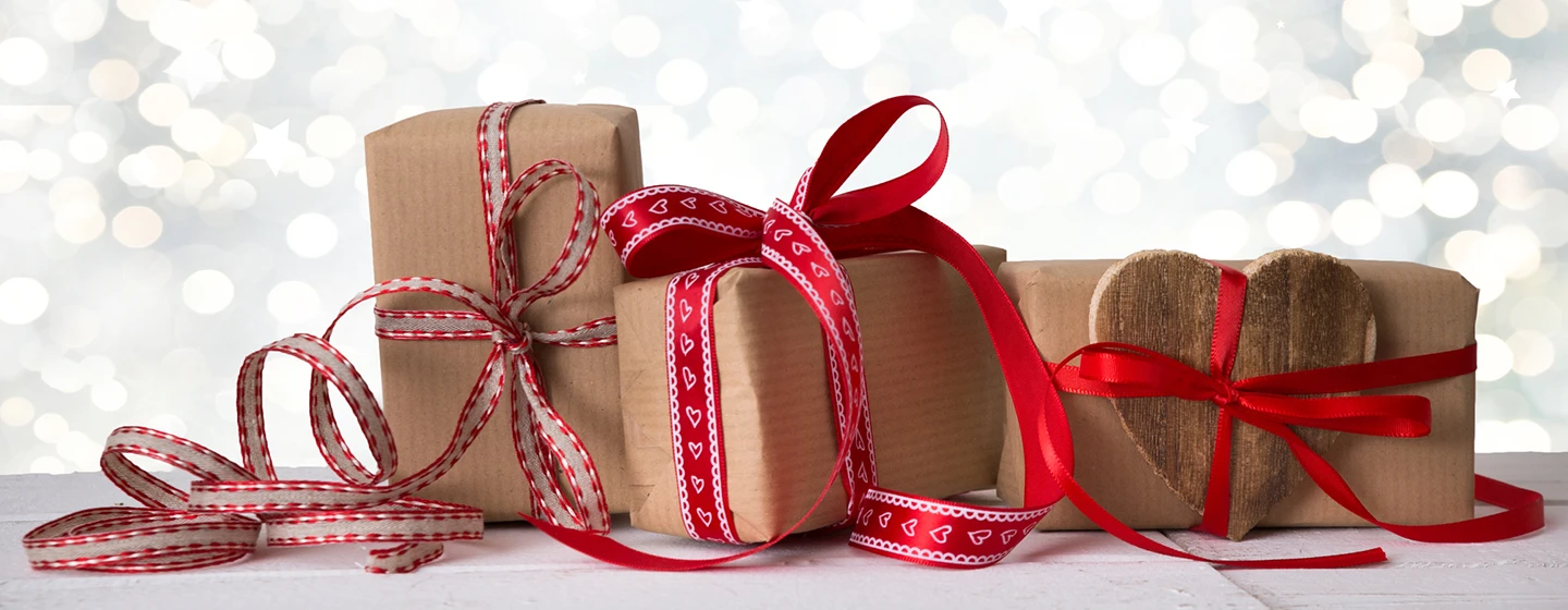 Drei in braunes Papier eingewickelte Geschenke mit roten und rot-weißen Bändern auf einer hellen Fläche, daneben ein herzförmiges Holzelement.